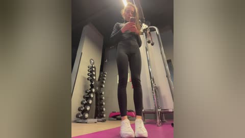 Media: Video of a young woman with long blonde hair, wearing a black sports bra and leggings, taking a selfie in a gym. Background includes weights, exercise equipment, and a pink mat.