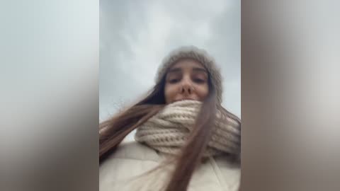 Media: Video of a young woman with long brown hair, wearing a thick cream-colored scarf and a furry hat, standing in a blurred, overcast outdoor setting.