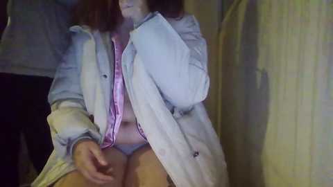 Media: Video of a woman with light skin and curly brown hair, sitting with legs crossed. She wears a light denim jacket over a pink blouse, revealing her white panties. Background includes a yellow curtain and a dimly lit room.