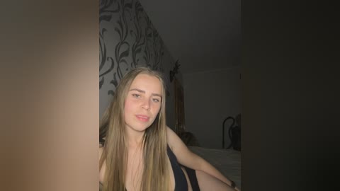 Media: Video of a young, fair-skinned woman with long, straight blonde hair, wearing a black tank top, leaning against a gray wall with ornate, black floral patterns.
