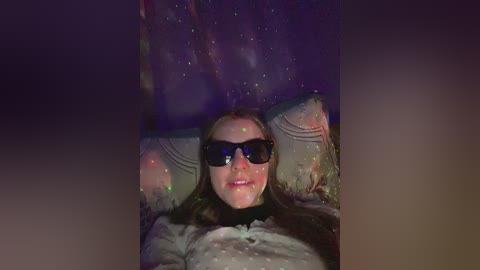 Video of a woman with long brown hair, wearing dark sunglasses, lying on a bed with a starry night sky projection.