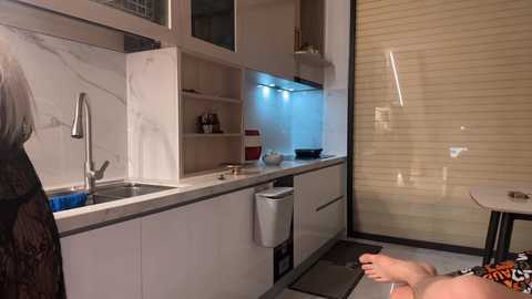 Media: Video of a modern kitchen with white cabinets, marble countertops, stainless steel sink, and a person in a striped shirt and black pants washing dishes. The background features a closed roll-up window.