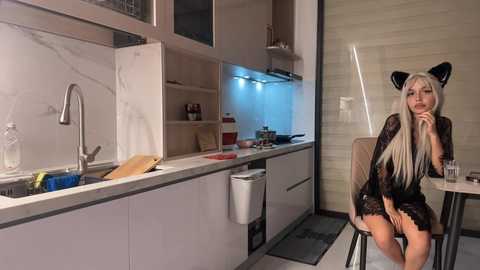 Media: Video of a modern kitchen with a blonde woman in a black lace dress and cat ears, sitting on a stool, with a counter and sink in the background.