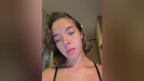 Media: Video of a young woman with light brown, wavy hair and fair skin, wearing a black spaghetti-strap top, looking contemplative in a dimly lit, blurry bathroom setting.