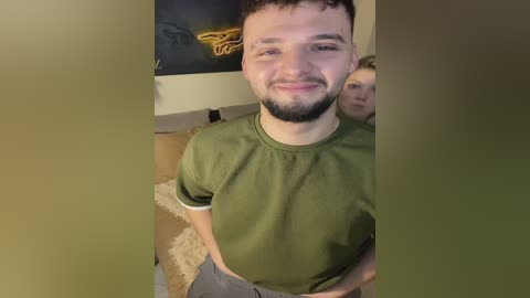 Media: Video of a young man with short brown hair, a beard, and a slight smile, wearing a green T-shirt, standing indoors with a blurred background featuring a wall-mounted art piece.