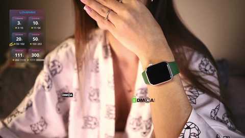 Media: Video of a woman with long brown hair, wearing a white robe with black panda patterns, and a green smartwatch, holding her hand to her face.