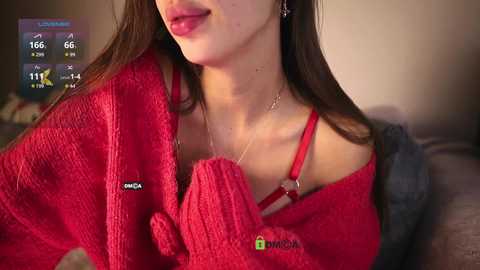 Media: Video of a young woman with fair skin, long brown hair, wearing a red knit sweater, a red bra strap showing, and a necklace. Background shows a blurred living room and a digital thermometer display.