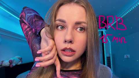 Media: Video of a young, light-skinned woman with long, straight blonde hair, wearing a pink and purple floral top. She has light makeup, green eyes, and pink lipstick, with a surprised expression. Background shows a dimly lit room with neon blue lighting, a bed, and a pink \"BBB FAN\" logo.