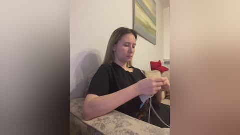 A video of a Caucasian woman with long brown hair, wearing a black shirt, sitting on a patterned couch, holding a syringe, in a beige room with a green painting on the wall.