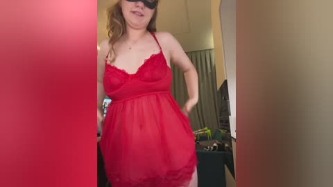 A video of a fair-skinned, curvy woman in a red lace lingerie dress, wearing a black mask, standing indoors with a partially blurred background.