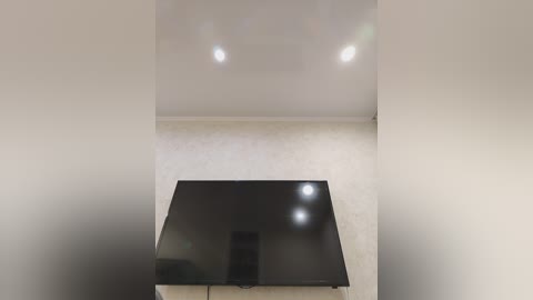 Video of a minimalist room with a large, flat-screen TV mounted on a beige wall. The room features smooth, light-colored walls and ceiling, with two recessed ceiling lights providing soft illumination.