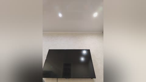 Video of a minimalist room with a sleek, black flat-screen TV mounted on a beige wall, centered on a white textured wall. Two small recessed ceiling lights provide soft illumination.
