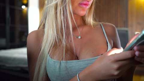 Media: Video of a blonde woman with long hair, wearing a light gray tank top that accentuates her large breasts, holding a phone. She has a bracelet on her right wrist. The background is blurred, showing a wooden wall and a bed.