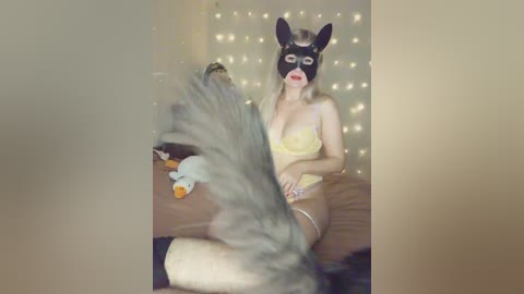 A video of a woman in a yellow lingerie set, wearing a black dog mask, sitting on a bed with a fluffy grey cat. The background is softly lit with fairy lights.