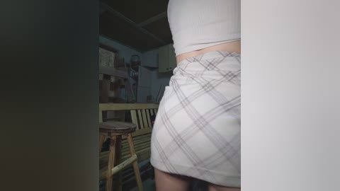 A video of a person wearing a plaid skirt and crop top, standing in a dimly lit, cluttered room with a wooden bench and bookshelves in the background.