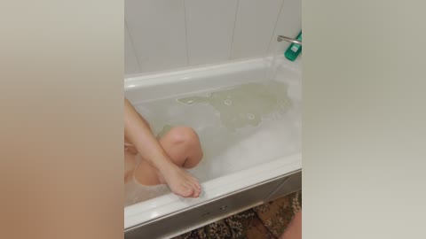 A video of a person with light skin and short hair, partially submerged in a bathtub filled with water and bubbles, leaning against the white-tiled wall.