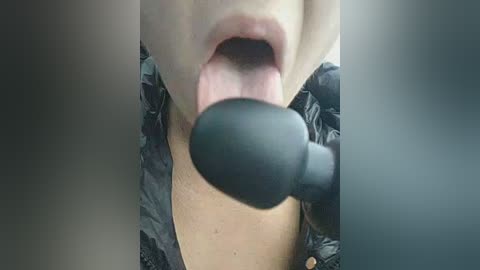 Video of a close-up of a woman's mouth open, tongue extended, with a black dildo near her lips. She wears a dark patterned jacket.