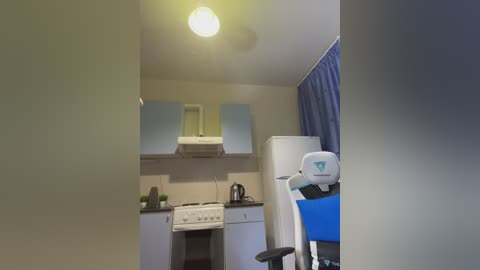 A video of a small, dimly lit kitchen with white cabinets, a stove, a fridge, and a gaming chair with a blue and white logo.