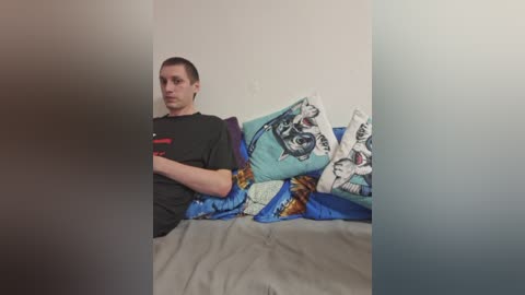 A video of a young man with short dark hair, wearing a black T-shirt, lying on a bed with a blue and green duvet cover featuring a cat design, surrounded by a blurred, beige room.