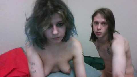 Media: Video of a nude, pale-skinned woman with shoulder-length, dark hair and a tattoo on her left arm, sitting on a bed with a red pillow. Behind her, a thin, pale-skinned man with shoulder-length brown hair, also nude, looks at the camera.