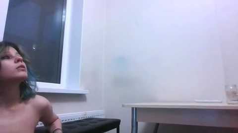 Media: Video of a nude woman with blue hair, standing against a white wall, near a window. A white table and a glass of water are visible in the background.