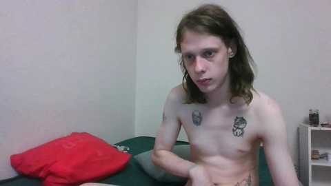 Media: Video of a skinny, shirtless white male with shoulder-length brown hair, wearing glasses, and a tattoo of a skull on his right pectoral. He sits on a green mattress in a dimly lit room with white walls, a red pillow, and a small white shelf in the background.