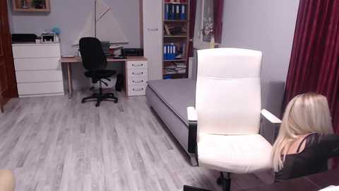 Media: Video of a modern, well-organized home office with a white leather chair facing a gray bed, light wooden flooring, a desk with a black chair, and bookshelves filled with binders and decor.