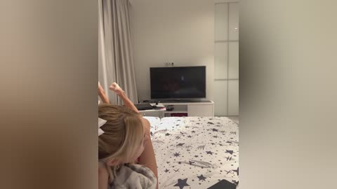 Media: Video of a blonde woman in bed, wearing a white top, with a TV on a white stand in the background. The room has a minimalistic, modern decor.