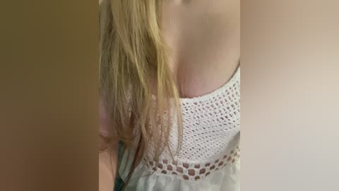 Video of a light-skinned woman with long, blonde hair, wearing a white, crochet top, partially covering her bare shoulders and cleavage. The background is a plain beige wall.
