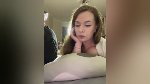 Media: A video of a young woman with light skin and long brown hair, wearing a white top, resting her head on her hand, looking contemplative.