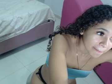 Video of a young Latina woman with curly black hair, light brown skin, wearing a blue strapless top and black bikini bottoms, sitting on a pink cushion on a tiled floor beside a bed with a white sheet.