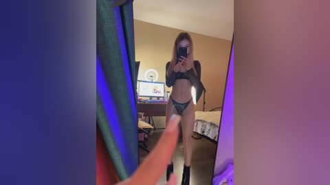 A video of a slender, light-skinned woman with long, straight hair in black lingerie and knee-high socks, taking a mirror selfie in a dimly lit bedroom with a bed and TV in the background.