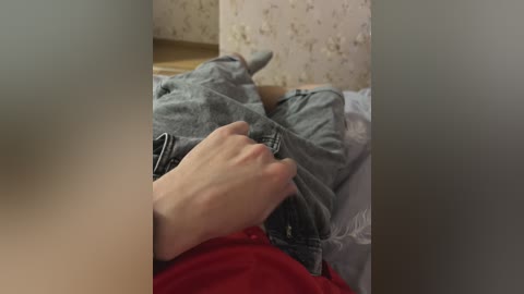 A video shows a person's hand in a red shirt lifting a light-colored shirt with blue jeans, lying on a bed with white sheets. The background features patterned wallpaper.