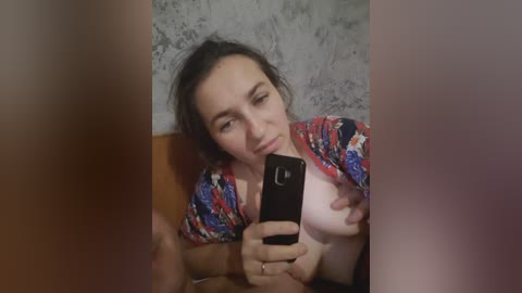 A video of a young woman with fair skin, dark hair, and a red floral top, taking a selfie with a black smartphone, partially obscured by another person's arm.