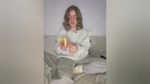 Video of a young woman with wavy brown hair, wearing a white sweatshirt and gray pants, eating a banana on a beige couch.