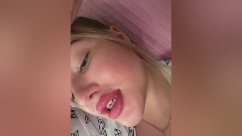 A video of a blonde woman with light skin, wearing red lipstick, lying on a bed with a white pillowcase featuring black geometric patterns. The background includes a pink blanket.