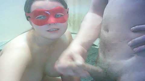 Video of a nude, fair-skinned woman with red mask and blue eyes, kneeling and grasping a flaccid, circumcised penis.