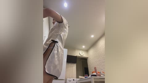 A video of a person, possibly male, in a white satin robe, leaning against a beige wall in a modern kitchen with white brick accents and a clock on the wall.