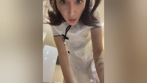 A video of an Asian woman with light skin and dark hair, wearing a shiny white nurse uniform, standing in a bathroom with beige walls and a white toilet.