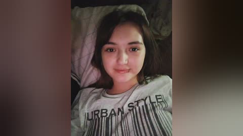 Media: A video of a young Asian woman with long black hair, lying on a bed, wearing a gray \"Urban Style\" T-shirt. She has a slight smile and is surrounded by dim lighting.