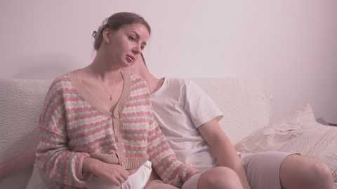 Video of a young, fair-skinned woman with brown hair in a messy bun, wearing a striped cardigan and white shirt, sitting on a beige couch, holding a white pillow, next to a man with short hair, in a softly lit room.