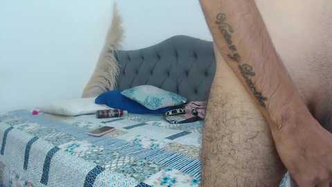 A video of a man's hairy leg and tattooed forearm, standing next to a gray upholstered bed with a quilted bedspread, pillows, and a feather pillow.