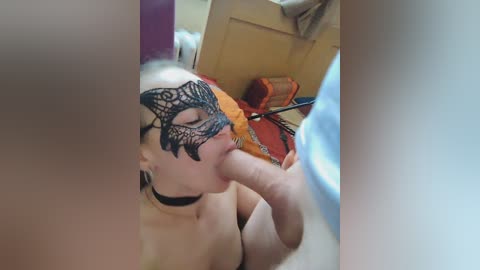 Video of a fair-skinned woman with blonde hair, wearing a black lace masquerade mask, performing oral sex on a man's erect penis. Background shows a wooden door and a colorful bag.