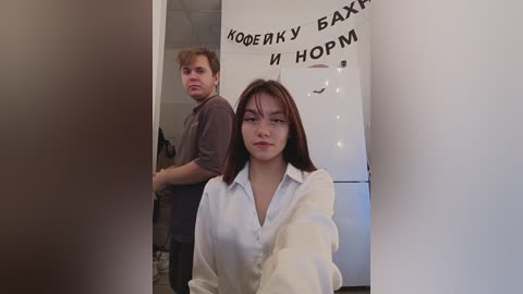 Media: Video of a young woman with long brown hair, wearing a white shirt, standing in a hallway. A man with short brown hair in a gray shirt stands behind her, both looking serious.