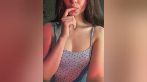 Video of a young woman with long, straight black hair, wearing a blue spaghetti-strap dress with a subtle pattern, standing indoors with her finger to her lips. Warm lighting casts a red glow on her face.