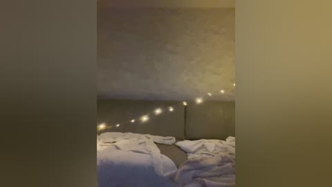 A dimly lit video of a bedroom with a white bed covered in messy white sheets, a dark headboard, and a string of small, warm yellow lights draped across the top.