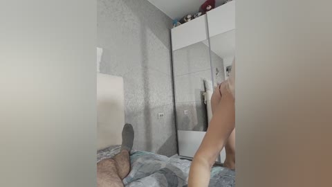 Media: A video of a modern, minimally decorated bedroom with a large mirror reflecting a person in the foreground, wearing grey socks. The room features a white bed with grey and white bedding, and a white closet with a few items on top.