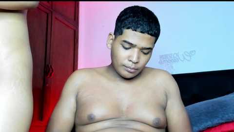 Video of a shirtless, young, fit, dark-skinned man with short black hair, sitting on a bed. Background features red and blue walls with a white pillow and a \"Daddy\" text.