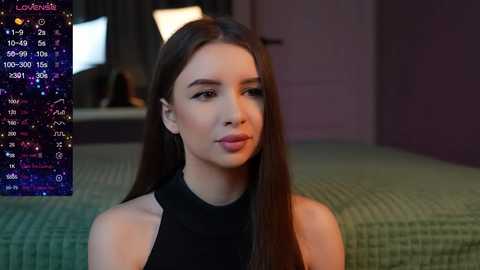 Media: Video of a young woman with long, straight dark hair, wearing a black sleeveless top, sitting on a green quilted bed in a dimly lit room. Overlay shows astrology symbols and text.