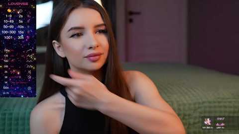 Media: Video of a young woman with long brown hair, wearing a black sleeveless top, pointing with her left hand, in a bedroom with a green quilted bedspread, purple walls, and a digital astrology screen showing \"Lover\" in the background.